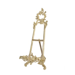 May include: A gold-colored decorative easel with ornate scrollwork. The easel has a three-legged design, with a decorative base and a detailed top. The frame is designed to hold a picture or artwork.