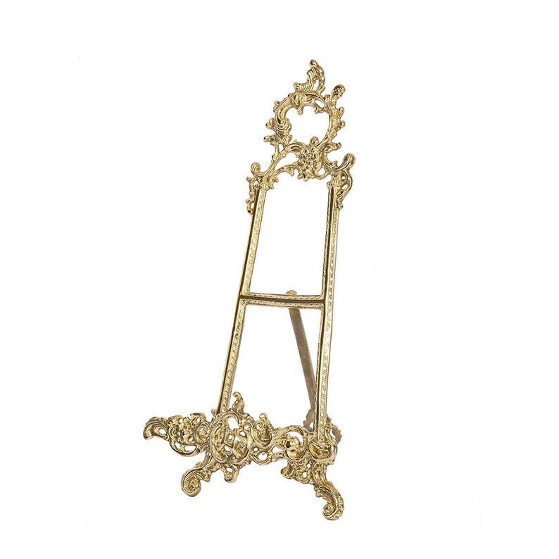 May include: A gold-colored decorative easel with ornate scrollwork. The easel has a three-legged design, with a decorative base and a detailed top. The frame is designed to hold a picture or artwork.