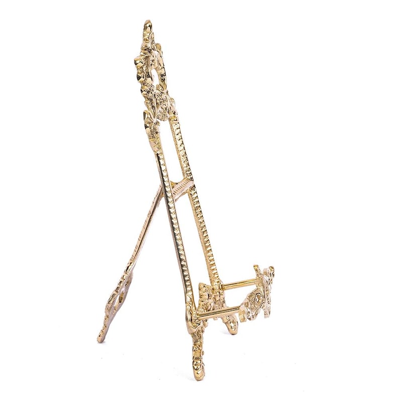 May include: A gold metal easel stand with ornate details. The stand has a decorative top and three legs.