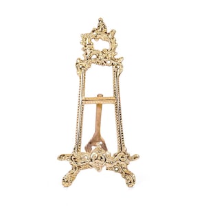 May include: A gold metal easel with an ornate design. The easel has a rectangular frame and three legs.