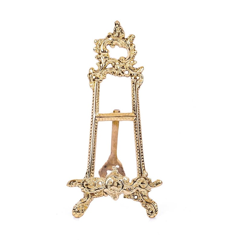 May include: A gold metal easel with an ornate design. The easel has a rectangular frame and three legs.