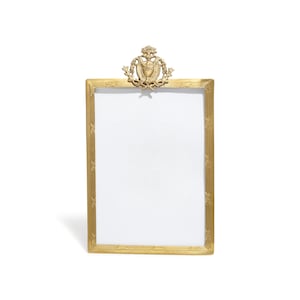 May include: A gold-toned picture frame with an ornate design. The frame has a decorative crest at the top and a rectangular opening for a photo.