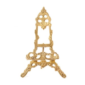 May include: A decorative gold-coloured easel with ornate detailing. The easel has a three-legged base and a detailed frame, suitable for displaying artwork or small items. The design includes floral and scrollwork patterns.