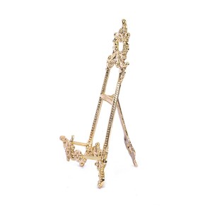 May include: A gold metal easel with ornate details. The easel is designed to hold a picture or other artwork.