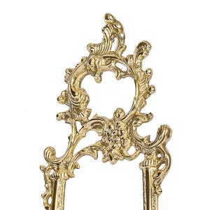 May include: Ornate gold-colored metal decorative stand with a floral and scroll design. The stand has a central open circle and two vertical supports. The decorative elements are detailed and symmetrical.