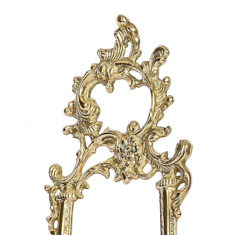 May include: Ornate gold-colored metal decorative stand with a floral and scroll design. The stand has a central open circle and two vertical supports. The decorative elements are detailed and symmetrical.