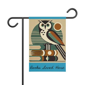 May include: A garden flag featuring a stylized owl perched atop a stack of books. The owl has a white body, black and green eyes, and brown and teal accents. The flag has the text "Books Loved Here" in a teal banner.
