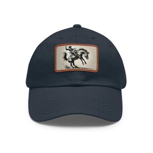 Può includere: Cappellino da baseball blu navy con una toppa rettangolare con un'illustrazione in bianco e nero di un cowboy su un cavallo selvaggio. La toppa ha un bordo marrone. Il cappellino ha una visiera curva.