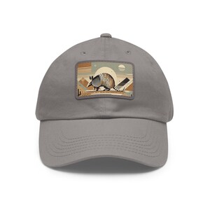 May include: A gray baseball cap featuring a rectangular patch with an armadillo illustration. The armadillo has a pattern resembling the American flag. The background includes a desert landscape with a sun and geometric shapes. The cap is made of a soft fabric.