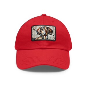 May include: A bright red baseball cap featuring a geometric dachshund design on a rectangular patch. The patch showcases the dog's face with brown, black, and white geometric shapes. The cap has a curved brim and an adjustable strap.