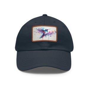 Puede incluir: Gorra de béisbol azul marino con un parche rectangular con un diseño de colibrí en acuarela. El colibrí está representado con tonos azules, morados y rojos, sobre un fondo blanco con salpicaduras de colores.