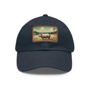 Puede incluir: Una gorra de béisbol azul marino con un parche rectangular en la parte delantera. El parche muestra una ilustración colorida de un cerdo de pie en un campo de hierba con un molino de viento al fondo bajo un cielo azul.