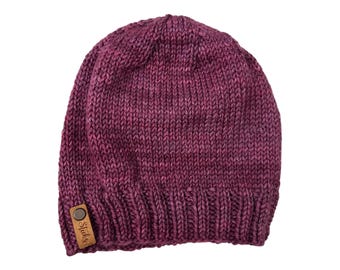 May include: A knitted beanie in shades of burgundy and purple. The hat features a ribbed brim and a small, brown leather tag with the text "Stitch's". The hat is made of a thick, textured yarn.