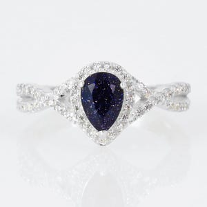 Op de afbeelding: Zilveren ring met een peervormige, donkerblauwe edelsteen, omringd door een halo van kleine, heldere stenen. De band heeft een gedraaid ontwerp. De ring staat tegen een witte achtergrond. De donkerblauwe edelsteen heeft een sprankelend effect.