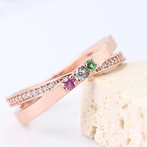 Puede incluir: Anillo de oro rosa con un diseño entrecruzado. Una banda está incrustada con pequeñas piedras transparentes. El anillo presenta un rubí, una piedra transparente y una esmeralda. El anillo se muestra sobre una superficie clara y texturizada.