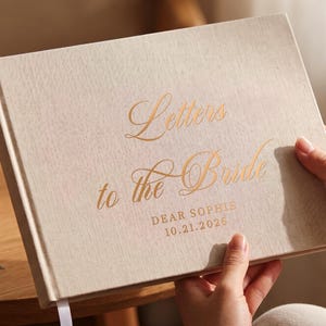 Puede incluir: Un libro de visitas con cubierta de tela beige y letras doradas que dicen Letters to the Bride, DEAR SOPHIE y 10.21.2026. El libro rectangular de tapa dura incluye un marcapáginas de cinta blanca que sobresale por el borde inferior.