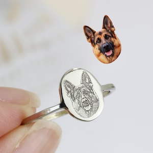 Op de afbeelding: Een zilveren ring met een gegraveerd portret van een Duitse herdershond op een ovale zetting. De ring wordt omhoog gehouden, met een aparte afbeelding van de kop van een Duitse herdershond op de achtergrond. De ring is een sieraad met een huisdierthema.
