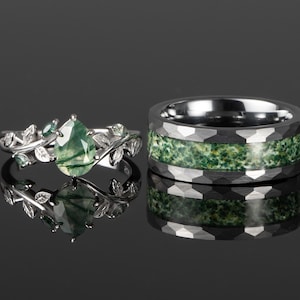 Könnte beinhalten: Zwei Ringe werden gezeigt. Einer ist ein silberner Ring mit einem birnenförmigen grünen Stein und Blattakzenten. Der andere ist ein dunkelgrauer Ring mit einem grün gesprenkelten Inlay. Beide Ringe befinden sich auf einer reflektierenden Oberfläche.