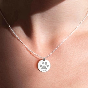 Puede incluir: Un collar plateado con un colgante redondo con un diseño de huella de pata. El colgante cuelga de una cadena fina. El collar se lleva alrededor del cuello.
