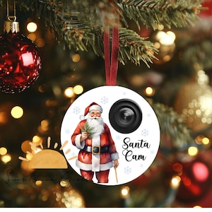 Puede incluir: Adorno navideño redondo con Papá Noel con traje rojo, sosteniendo una ramita verde. Una lente de cámara negra y las palabras "Santa Cam" también están en el adorno. El adorno cuelga de una cinta roja.