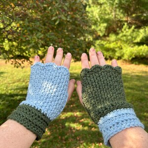 May include: A pair of handmade fingerless gloves. One glove is light blue with a dark green cuff, and the other is dark green with a light blue cuff. The gloves are crocheted and have a scalloped edge.