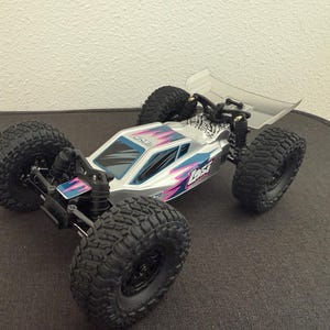 May include: A silver and black remote control car with large, black, knobby tires. The car has a clear spoiler and is decorated with pink and blue accents. The word "Lost" is visible on the side of the car.