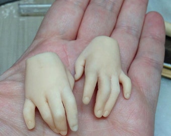 May include: Two small, pale-colored doll hands, one with a detached forearm, are held in a person's open palm. The hands are detailed with sculpted fingers and nails, suggesting a miniature craft or art project.