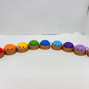 May include: A row of nine colourful dumpling keychains, each featuring a smiling face with rosy cheeks. The dumplings are red, orange, yellow, green, dark blue, light blue, purple, lavender, and pink, each resting in a small brown steamer basket.