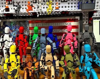 Op de afbeelding: Een verzameling veelkleurige, beweegbare robotactiefiguren in verschillende houdingen. De figuren staan voor een bord met de tekst I PLEDGE ALLEGIANCE TO THE FLAG OF THE UNITED STATES OF AMERICA. De robots zijn rood, blauw, groen, geel, roze, wit, zwart en beige.