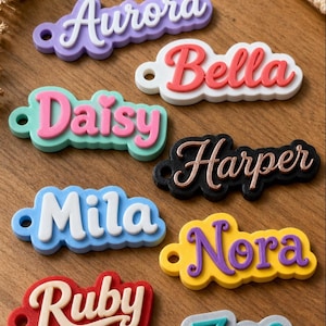 May include: A collection of custom name tags featuring raised lettering in various colors including purple, white, red, pink, black, blue, yellow, and gray. Each tag has a small circular hole on the left side for attachment and displays a unique name.