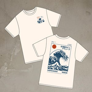 Może przedstawiać: Dwie kremowe koszulki. Jedna koszulka ma małą niebiesko-pomarańczową grafikę z napisem "Tokyo Japan". Druga koszulka ma dużą niebieską grafikę z falami z napisem "Hokkaido Wave" i japońskimi znakami.