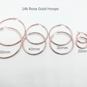 May include: A collection of 14k rose gold hoop earrings in various sizes. The sizes are 55mm, 40mm, 30mm, 25mm, and 15mm. The earrings are displayed on a white surface. The text "14k Rose Gold Hoops" is at the top.