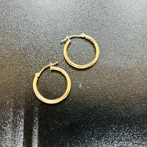 May include: A pair of gold hoop earrings. The earrings are circular and have a simple, classic design. The hoops are made of a shiny, gold-colored metal. The earrings are displayed on a dark, textured surface.