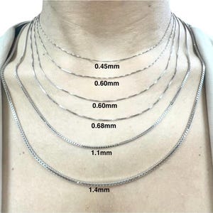 May include: A collection of silver necklaces of varying thicknesses, displayed on a person's neck. The necklaces range from 0.45mm to 1.4mm in width, with the measurements clearly labeled. The chains have different styles, including box and snake chains.