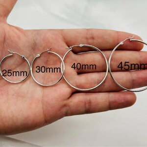 May include: Four silver hoop earrings in various sizes: 25mm, 30mm, 40mm, and 45mm. The earrings are displayed on a hand, showcasing their different diameters. The hoops are simple and classic, with a shiny finish.