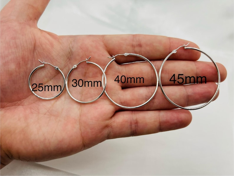 May include: Four silver hoop earrings in various sizes: 25mm, 30mm, 40mm, and 45mm. The earrings are displayed on a hand, showcasing their different diameters. The hoops are simple and classic, with a shiny finish.