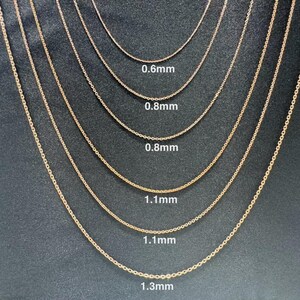 May include: A close-up of seven gold chains of different thicknesses, laid out in a row on a dark gray surface. The chains are labeled with their thickness in millimeters: 0.6mm, 0.8mm, 0.8mm, 1.1mm, 1.1mm, and 1.3mm.