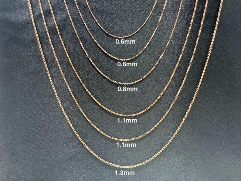 May include: A close-up of seven gold chains of different thicknesses, laid out in a row on a dark gray surface. The chains are labeled with their thickness in millimeters: 0.6mm, 0.8mm, 0.8mm, 1.1mm, 1.1mm, and 1.3mm.
