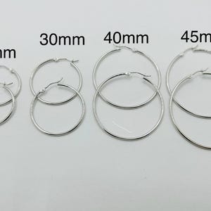 May include: A collection of silver hoop earrings in various sizes: 25mm, 30mm, 40mm, and 45mm. The earrings are displayed on a white background, showcasing their simple, elegant design. The sizes are clearly labeled above each pair.