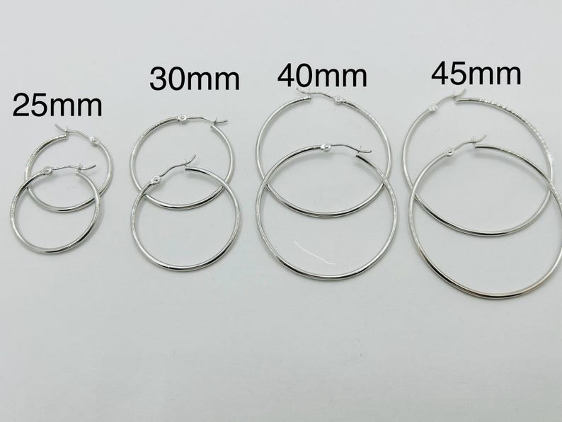 May include: A collection of silver hoop earrings in various sizes: 25mm, 30mm, 40mm, and 45mm. The earrings are displayed on a white background, showcasing their simple, elegant design. The sizes are clearly labeled above each pair.