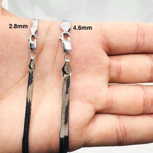 May include: Two silver herringbone chain necklaces, 2.8mm and 4.6mm wide, are displayed against a hand. Each chain has a lobster clasp closure. The chains are flat and have a reflective, metallic appearance.