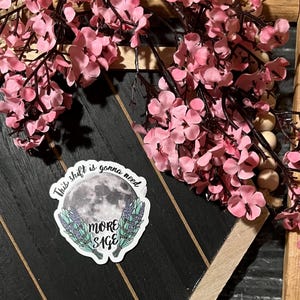 May include: A sticker with the text "This shift is gonna need MORE SAGE" featuring a moon and lavender sprigs, placed on a black and wood-toned surface. Pink cherry blossom branches with dark stems and wooden beads are arranged around the sticker.