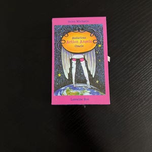 Puede incluir: Una caja rosa con el título "Audacious Action Angels Oracle" y los nombres de los autores "Helen Michaels" y "Lorraine Roe" impresos en la parte delantera. La caja presenta una ilustración de un ángel con alas de pie sobre un globo terráqueo.
