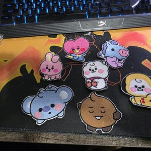 May include: A collection of embroidered patches featuring cartoon characters in various colors. The patches include a pink bunny, a blue koala, a brown bear, and other cute designs. They are arranged on a patterned surface near a keyboard.