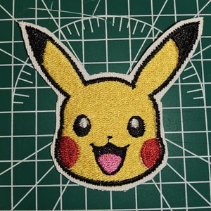 May include: An embroidered patch featuring the yellow head of a cartoon character with long ears tipped in black, round red cheeks, black eyes with white highlights, a small nose, and an open mouth showing a pink tongue, outlined in white thread.