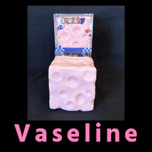 May include: A light pink, cube-shaped squishy toy with circular indentations resembling Swiss cheese, positioned in front of its clear plastic packaging. The packaging features the word Squishy in blue text with colorful geometric shapes.