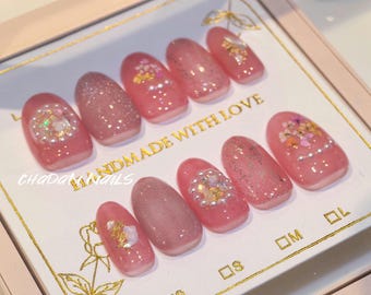 May include: A set of ten pink press-on nails with various designs. Some nails feature glitter, floral accents, and pearl embellishments. The nails are arranged on a white surface with gold text that reads "HANDMADE WITH LOVE".