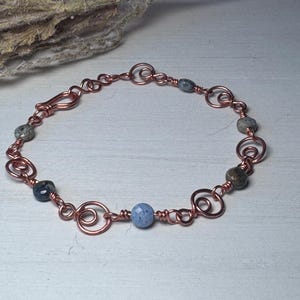 May include: A copper wire bracelet featuring a series of interconnected hand-formed wire spirals and small faceted beads in shades of blue, gray, and brown. The bracelet includes a hook-and-eye clasp mechanism made from the same copper wire.