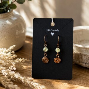 May include: A pair of dangle earrings featuring copper-colored wire spirals and small, pale green translucent beads. The earrings are attached to a black rectangular display card with the word Handmade and a small white heart icon printed on it.