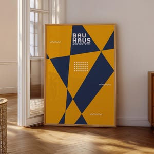 May include: A rectangular wall art print in a light wood frame featuring a geometric design of mustard yellow and dark blue triangles. The text BAU HAUS MODERNISTIC is printed in white at the top, accompanied by a grid of white dots and small zigzag lines.